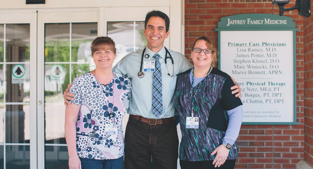 Three healthcare professionals from Jaffrey Family Medicine standing together and smiling in front of their clinic, showcasing their dedication to patient care.