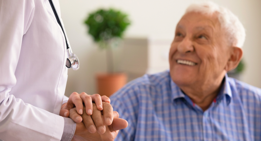 A smiling elderly man in a plaid shirt holds hands with a healthcare provider wearing a white coat and a stethoscope. The elderly man looks up at the provider with a happy and trusting expression, conveying a sense of comfort and reassurance in the care he is receiving. The background is softly blurred with a potted plant in the distance.