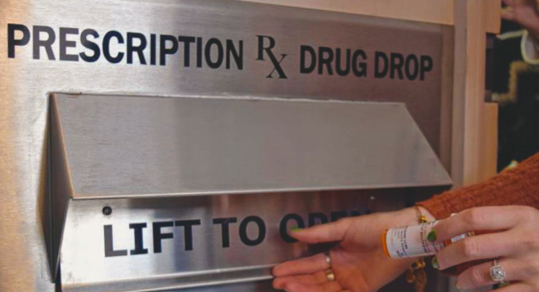 A close-up of a hand depositing a prescription bottle into a new prescription drug drop box labeled 'Prescription RX Drug Drop' at the Peterborough Police Department