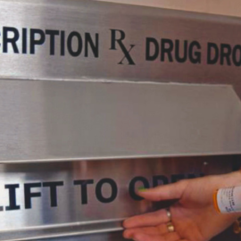 A close-up of a hand depositing a prescription bottle into a new prescription drug drop box labeled 'Prescription RX Drug Drop' at the Peterborough Police Department