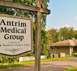 A large wooden sign reading 'Antrim Medical Group, A Service of Monadnock Community Hospital' stands in front of a single-story building surrounded by trees. The building has a light-colored exterior and is located in a serene, green setting