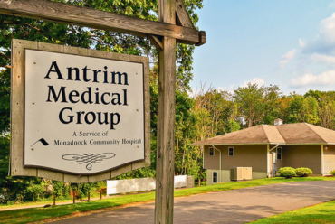 A large wooden sign reading 'Antrim Medical Group, A Service of Monadnock Community Hospital' stands in front of a single-story building surrounded by trees. The building has a light-colored exterior and is located in a serene, green setting