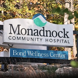 A sign for Monadnock Community Hospital with the Bond Wellness Center displayed underneath. The sign is surrounded by trees with autumn foliage, creating a vibrant backdrop