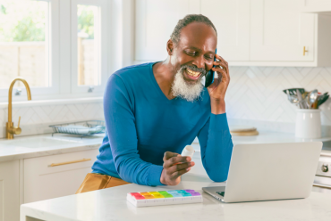 A smiling patient on the phone, holding a prescription bottle with his pill organizer and laptop in front of him