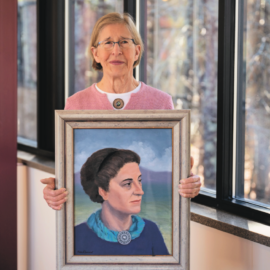 Karen Clement stands holding a framed portrait of her grandmother, Margaret Adams Clement, in a bright hallway at Monadnock Community Hospital. Margaret, depicted in the painting with a serene expression and blue scarf, was instrumental in founding the hospital over a century ago. The image symbolizes the Clement family's long-standing commitment to healthcare and community service.