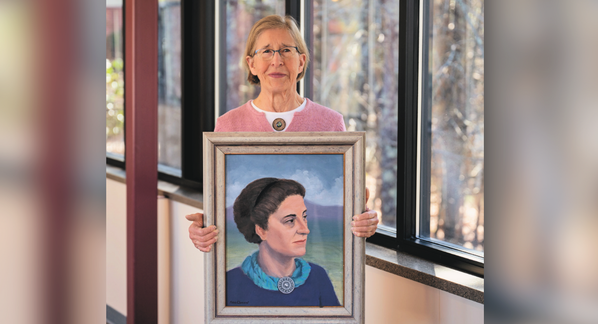 Karen Clement stands holding a framed portrait of her grandmother, Margaret Adams Clement, in a bright hallway at Monadnock Community Hospital. Margaret, depicted in the painting with a serene expression and blue scarf, was instrumental in founding the hospital over a century ago. The image symbolizes the Clement family's long-standing commitment to healthcare and community service.