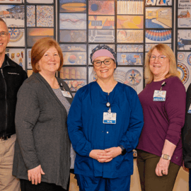 A group of Monadnock Community Hospital employees standing in front of a colorful mural. From left to right: Charles Trimble, wearing a black security vest and khaki pants; Cathy Maki, in a gray cardigan and black top; Pia Benet, dressed in blue scrubs with a headband; Karen Aucella, wearing a purple top and olive pants with a Rehab badge; and Jon Gullage, in a black jacket and blue scrub pants. The mural behind them is composed of various intricate patterns and images, representing the hospital's vibrant community and commitment to care.
