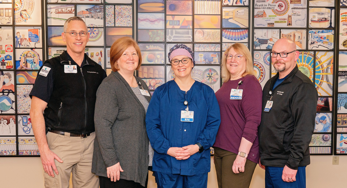 A group of Monadnock Community Hospital employees standing in front of a colorful mural. From left to right: Charles Trimble, wearing a black security vest and khaki pants; Cathy Maki, in a gray cardigan and black top; Pia Benet, dressed in blue scrubs with a headband; Karen Aucella, wearing a purple top and olive pants with a Rehab badge; and Jon Gullage, in a black jacket and blue scrub pants. The mural behind them is composed of various intricate patterns and images, representing the hospital's vibrant community and commitment to care.