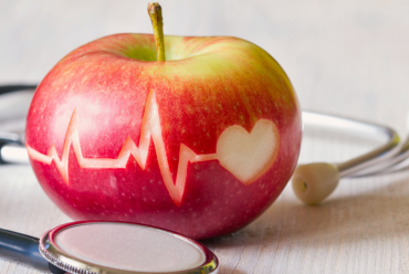 A red apple with a heart and a heartbeat line carved out of it and a stethoscope wrapped around it