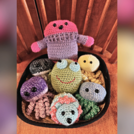 A collection of handmade crocheted stuffed animals, including a pink and purple figure with arms raised, a green frog with large blue eyes, a yellow round creature, and various other small, colorful animals with black button eyes, all arranged in a basket on a wooden chair.