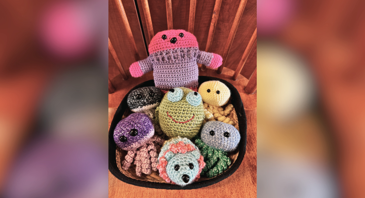 A collection of handmade crocheted stuffed animals, including a pink and purple figure with arms raised, a green frog with large blue eyes, a yellow round creature, and various other small, colorful animals with black button eyes, all arranged in a basket on a wooden chair.