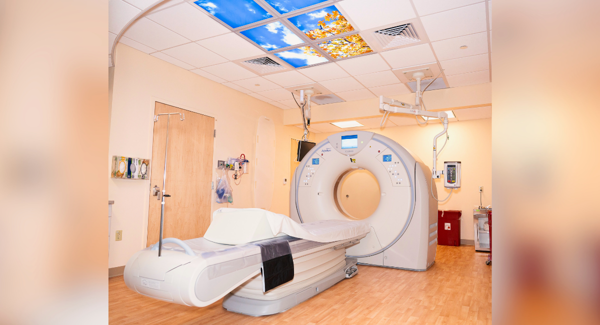 A modern radiology suite at Monadnock Community Hospital featuring a Toshiba 80-slice high-speed helical CT scanner. The CT scanner has a sleek, circular design with a comfortable patient table in front. The room is brightly lit, with warm beige walls and wood-patterned flooring, creating a welcoming atmosphere. Above the scanner, decorative ceiling panels depict a serene outdoor scene with blue skies and golden autumn leaves, adding a calming touch to the clinical environment. Medical equipment and monitors are visible around the room, highlighting the advanced technology used for diagnostic imaging.