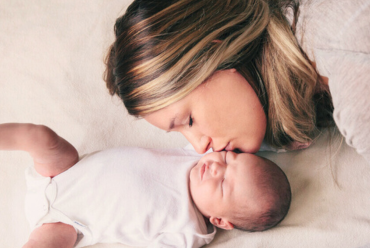 A mother with brown hair kisses her newborn in a white onesie