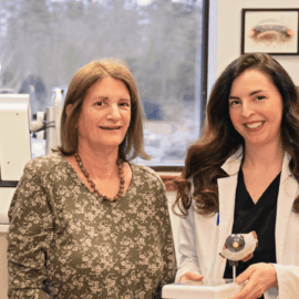 Dr. Heather Bartels and Patricia Shuster sit together in an examination room at Monadnock Community Hospital’s Medical Eye Center. Dr. Bartels, in a white coat, holds an eye model, while Patricia, wearing a floral dress, smiles beside her. Advanced medical equipment is visible in the background, highlighting the state-of-the-art facilities at the center.