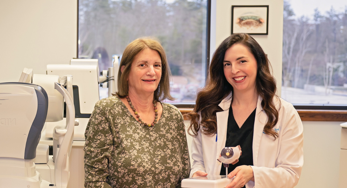 Dr. Heather Bartels and Patricia Shuster sit together in an examination room at Monadnock Community Hospital’s Medical Eye Center. Dr. Bartels, in a white coat, holds an eye model, while Patricia, wearing a floral dress, smiles beside her. Advanced medical equipment is visible in the background, highlighting the state-of-the-art facilities at the center.