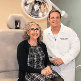Dr. Brian Kwitkin, a board-certified general surgeon, stands beside Hiyam Makarios in an examination room at Monadnock Community Hospital. Dr. Kwitkin is wearing a white coat and smiling, while Hiyam, sitting on the examination chair, wears a black-and-white patterned dress and glasses. They are surrounded by medical equipment, highlighting the hospital’s commitment to advanced surgical care.