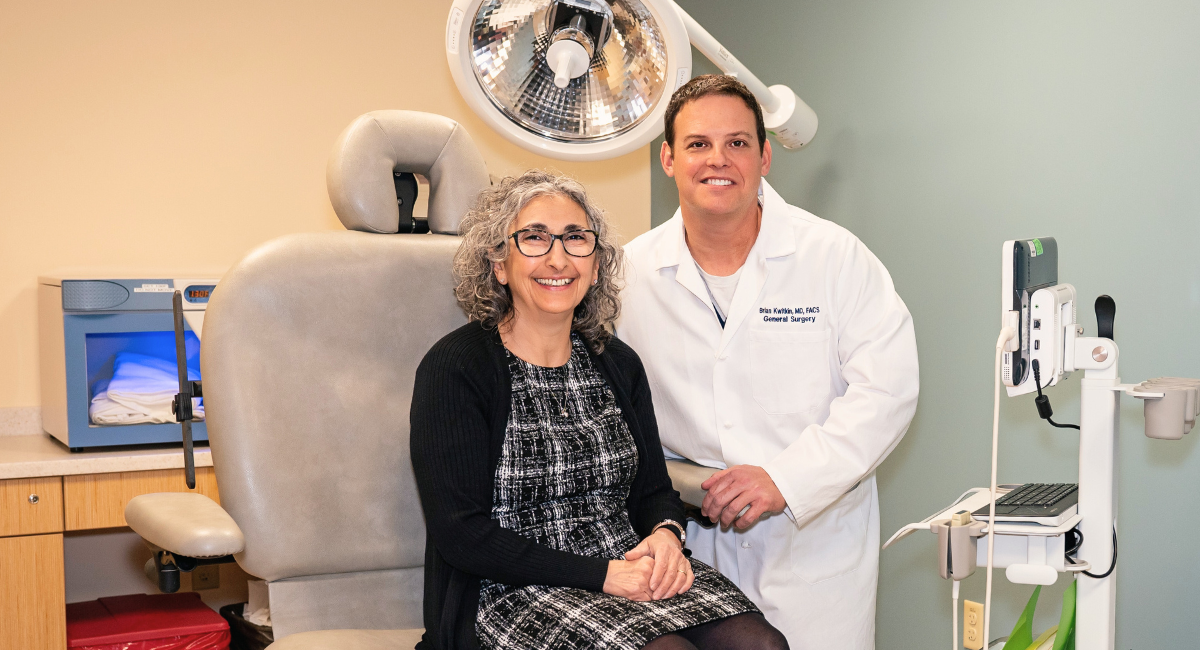 Dr. Brian Kwitkin, a board-certified general surgeon, stands beside Hiyam Makarios in an examination room at Monadnock Community Hospital. Dr. Kwitkin is wearing a white coat and smiling, while Hiyam, sitting on the examination chair, wears a black-and-white patterned dress and glasses. They are surrounded by medical equipment, highlighting the hospital’s commitment to advanced surgical care.