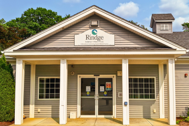 The front of Rindge Family Practice, a single-story building with a light gray exterior. A large white sign with the practice's name and the Monadnock Community Hospital logo is mounted above the entrance, flanked by white pillars.