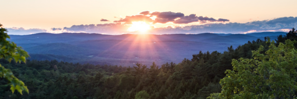 A purple sunset with scattered clouds in the sky, mountains in the distance, and evergreens in the foreground