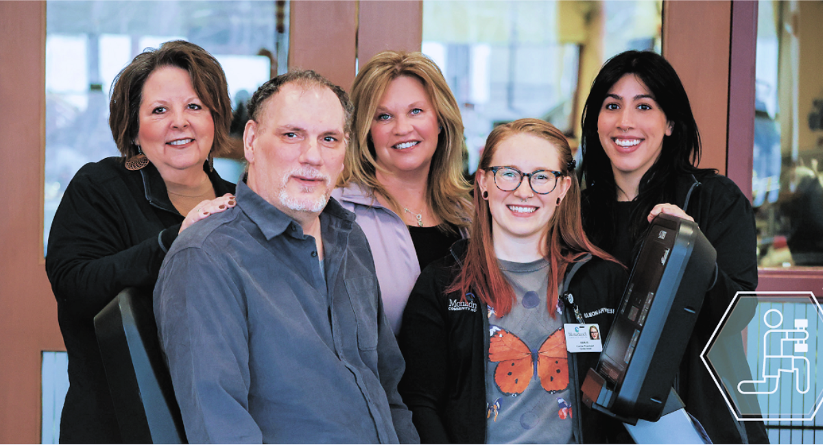 A group of Monadnock Community Hospital staff and patient Bruce Barsalou pose together in a bright, welcoming environment. From left to right: Essy Moverman, RRT, RCP, CTTS, Outpatient Pulmonary Coordinator, stands smiling with her hand on Bruce's shoulder. Bruce Barsalou, the patient, sits in a chair, smiling and relaxed. Dawne Beamer, a Respiratory Therapist, stands next to Bruce with a friendly expression. Kiara Burek, an Exercise Physiologist, stands beside Dawne, wearing glasses and a butterfly-themed shirt. Gabby Trust, also an Exercise Physiologist, stands on the far right, holding exercise equipment and smiling. The group is situated in front of large windows that let in natural light, symbolizing hope and recovery.