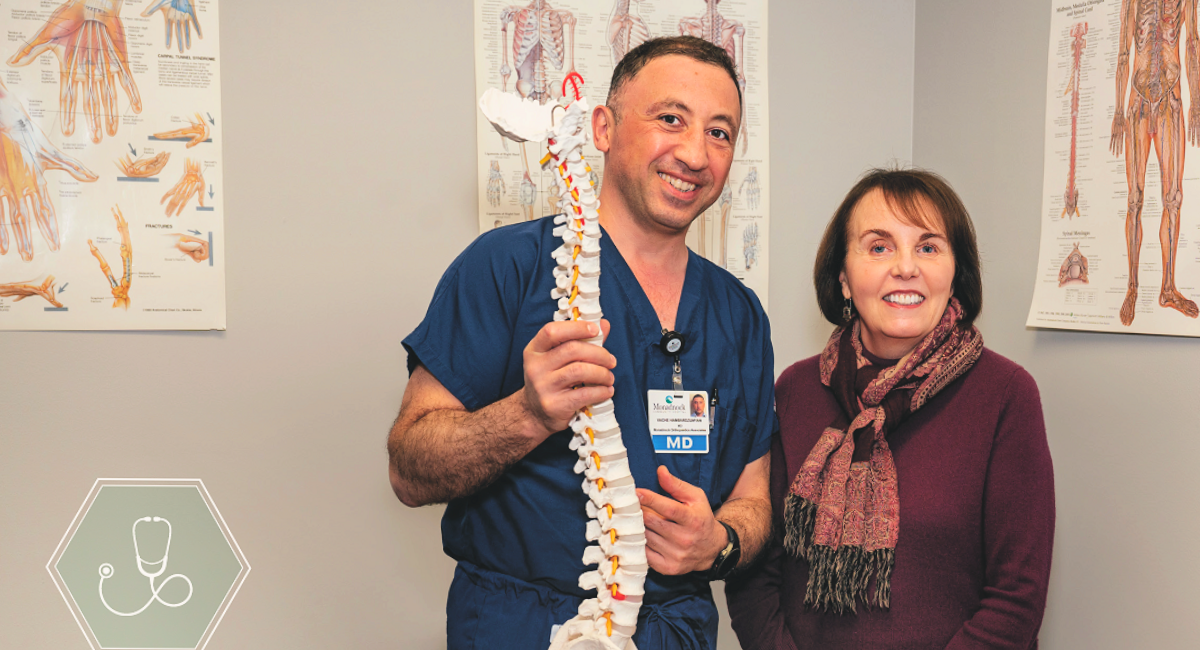 Dr. Vache Hambardzumyan, a physician at Monadnock Orthopaedic Associates, stands next to Louise DelPapa in a medical examination room. Dr. Hambardzumyan, dressed in blue scrubs, is holding a model of the human spine and smiling at the camera. Louise, wearing a maroon sweater and a patterned scarf, stands beside him with a smile. The background features anatomical posters of the human body, highlighting the clinical setting.