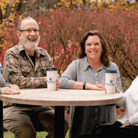 A group of Monadnock Community Hospital volunteers enjoying a moment together at an outdoor table. From left to right: Barbara Pendleton, wearing a green and blue sweater and a volunteer badge; Dewey Clark, in a plaid shirt and glasses, smiling with a Monadnock-branded coffee cup in front of him; Toni Gildone, the Volunteer Coordinator, wearing a gray button-up shirt and holding a Monadnock-branded coffee cup; and Connie Boyd, seated in a white sweater with a volunteer badge, smiling warmly at the camera. Behind them is a backdrop of autumn foliage with vibrant red and orange leaves, creating a welcoming and cheerful atmosphere.