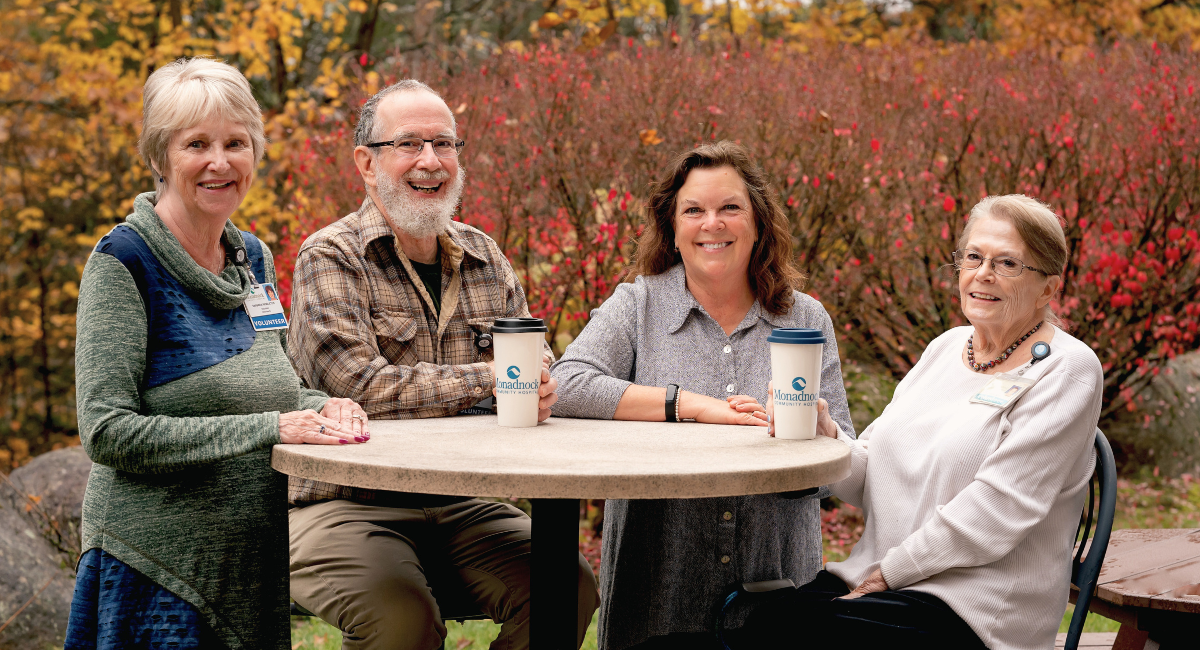 A group of Monadnock Community Hospital volunteers enjoying a moment together at an outdoor table. From left to right: Barbara Pendleton, wearing a green and blue sweater and a volunteer badge; Dewey Clark, in a plaid shirt and glasses, smiling with a Monadnock-branded coffee cup in front of him; Toni Gildone, the Volunteer Coordinator, wearing a gray button-up shirt and holding a Monadnock-branded coffee cup; and Connie Boyd, seated in a white sweater with a volunteer badge, smiling warmly at the camera. Behind them is a backdrop of autumn foliage with vibrant red and orange leaves, creating a welcoming and cheerful atmosphere.