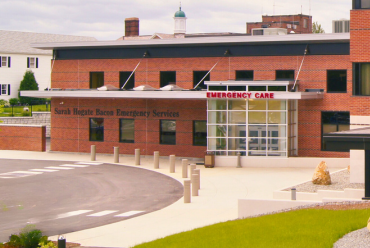 The entrance to the Sarah Hogate Bacon Emergency Services at Monadnock Community Hospital. The building has a modern brick facade with a glass entrance labeled "Emergency Care." A driveway leads directly to the entrance for easy access.