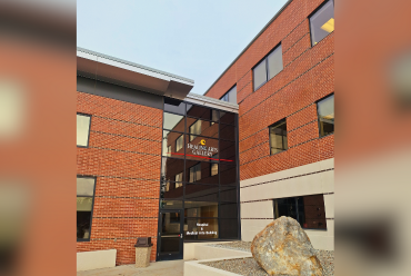 The entrance to the Healing Arts Gallery at Monadnock Community Hospital. The building features a brick exterior with large windows and a glass doorway prominently displaying the "Healing Arts Gallery" signage. A large decorative boulder sits in the landscaped area near the entrance.