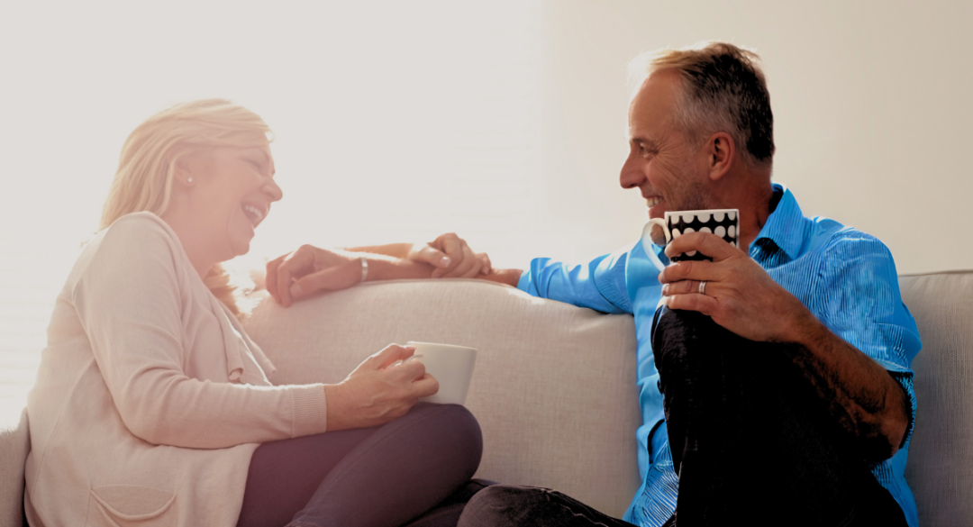 A middle-aged couple sitting on a couch, sharing a lighthearted conversation over coffee, symbolizing the importance of emotional support and social connection in managing chronic conditions like diabetes