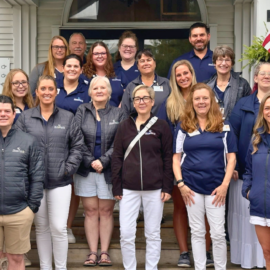 Group photo of Monadnock Community Hospital volunteers and staff standing on steps outside a building. Everyone is smiling, dressed in casual and branded attire, including jackets and polo shirts. The group includes men and women of various ages, with some wearing name badges and branded clothing featuring the hospital logo.