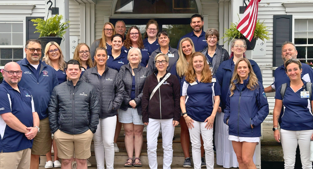 Group photo of Monadnock Community Hospital volunteers and staff standing on steps outside a building. Everyone is smiling, dressed in casual and branded attire, including jackets and polo shirts. The group includes men and women of various ages, with some wearing name badges and branded clothing featuring the hospital logo.