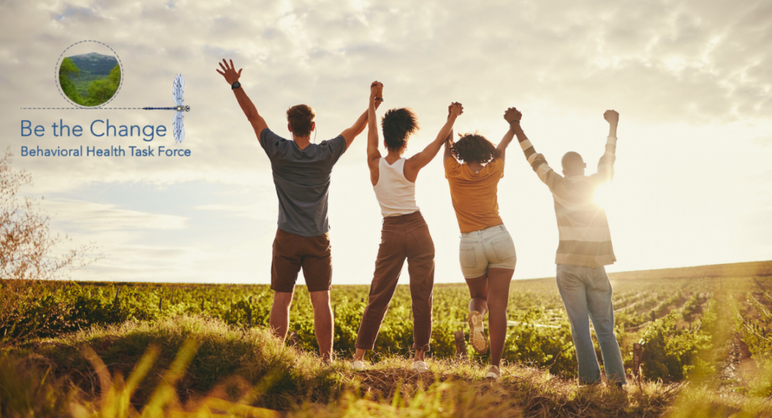 Four individuals standing together in a grassy field, facing away from the camera with their hands raised in unity as the sun sets behind them. The 'Be the Change Behavioral Health Task Force' logo is displayed on the left side, featuring a dragonfly and a landscape icon, symbolizing collective action and support for mental health