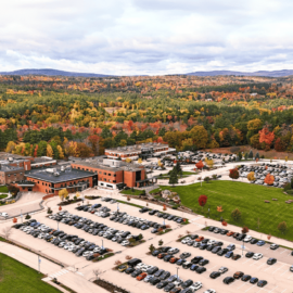 Aerial view of Monadnock Community Hospital, surrounded by vibrant autumn foliage, with parking lots in the foreground and rolling hills in the background. The scene captures the hospital buildings nestled in a colorful forest landscape, with clear skies and distant mountains visible on the horizon