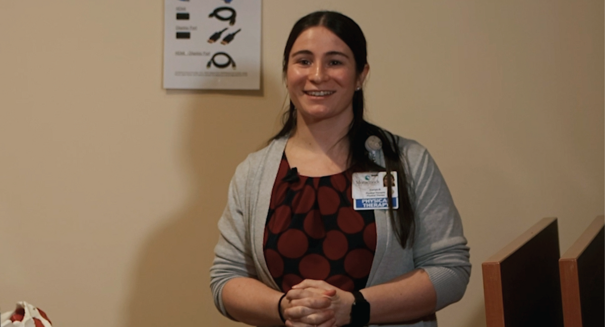 Corryn Nelson, PT, DPT, standing with hands clasped, smiling warmly during a seminar presentation. She is wearing a red and black patterned top under a gray cardigan. Behind her is a poster on the wall, and to the side, a table with a water bottle and anatomical model