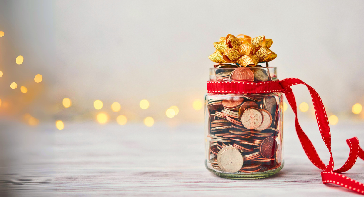 A glass jar filled with coins, wrapped with a red ribbon and topped with a gold bow, symbolizing charitable giving. The background features soft, golden lights, creating a warm and festive atmosphere