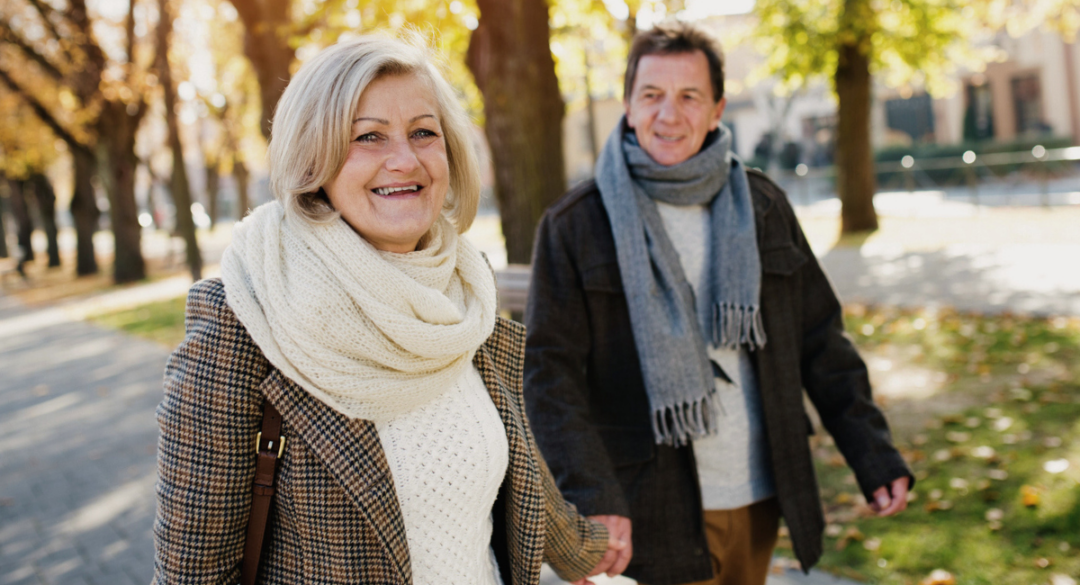 Smiling older woman in a plaid coat and scarf walking outdoors on a tree-lined path during autumn, holding hands with a man in a dark jacket and scarf, enjoying a sunny day