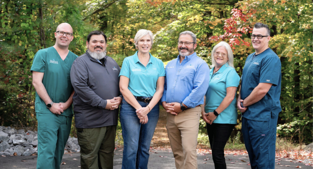 Group photo of the Monadnock Community Hospital Mobile Integrated Health (MIH) team. Six team members, dressed in various MCH uniforms, stand outdoors with a background of autumn foliage, smiling and looking at the camera. The team includes both clinical and support staff, reflecting a commitment to community healthcare