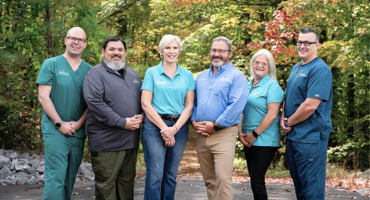 "Group photo of the Monadnock Community Hospital Mobile Integrated Health (MIH) team. Six team members, dressed in various MCH uniforms, stand outdoors with a background of autumn foliage, smiling and looking at the camera. The team includes both clinical and support staff, reflecting a commitment to community healthcare