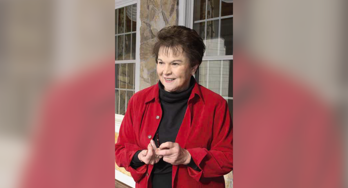 Photo of Pat Boyd wearing a red jacket and a black turtleneck, smiling warmly while gesturing with their hands, standing indoors with large windows and a stone wall in the background