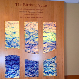 A wood-paneled dedication plaque for "The Birthing Suite," featuring colorful artistic panels and a recognition message for Corinne B. MacLennan-Kennedy in memory of her mother