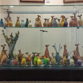 A collection of vibrant ceramic animals, including whimsical birds and animal figures, arranged on two shelves in a glass display case.