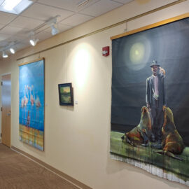 A hospital hallway gallery with various artworks, including vibrant paintings of a frog and flowers on one wall, and a man with dogs on the other. The artwork brings a touch of nature and color to the clinical setting.