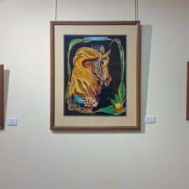 Three framed paintings displayed on a wall. On the left, a painting of a giraffe's head above water with a dark background; in the center, a large colorful depiction of a carousel horse framed with leaves and flowers; on the right, a painting of trees by a blue river in a forest scene