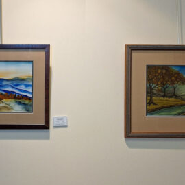 Two framed landscape paintings displayed side by side on a wall. The left painting shows mountain ranges in shades of blue and orange tones under a pale sky. The right painting features a countryside scene with autumnal trees beside a winding river under a bright sky