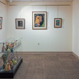 An alcove in the hospital gallery featuring a mix of framed artwork and a glass display case. The case contains a collection of small, colorful sculptures. Paintings on the wall depict various natural scenes and animals, enhancing the gallery's inviting atmosphere.