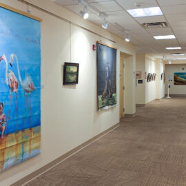 A hallway gallery with vibrant artworks, including a large flamingo painting on the left, smaller framed pieces along the right, and a seascape at the far end, all displayed in a well-lit space