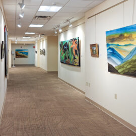 A long, well-lit hospital hallway showcasing a variety of colorful paintings on both sides. On the right side, a large landscape painting in blue, green, and yellow hues catches the eye. The hallway carpet is patterned and beige, contributing to a calm and welcoming atmosphere.