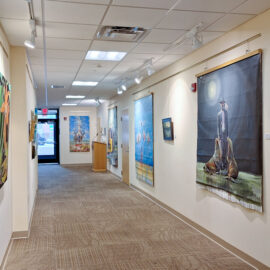 A hospital hallway gallery with various artworks, including vibrant paintings of a frog and flowers on one wall, and a man with walruses on the other. The artwork brings a touch of nature and color to the clinical setting.