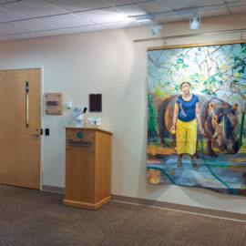 A gallery space featuring two large paintings: one of a woman with a hippo and another with abstract imagery of antelopes. A Monadnock Community Hospital podium stands between the artworks.